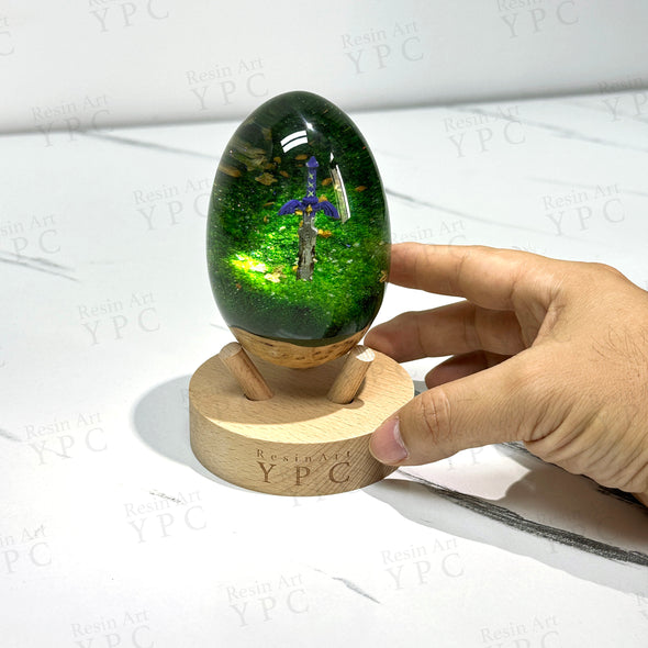 A resin and wood egg, larger than a goose egg, inside the egg is a green forest with a rusty master sword stuck in the middle, this egg comes with a wooden stand to display it. Inspired by The Legend of Zelda games